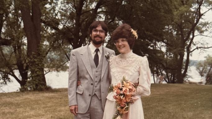 Pete and Nancy Torpey's wedding photo.  They are smiling broadly with their arms around each other's waists and standing on a grassy lawn with large lush trees and Irondequoit Bay behind them.  He is wearing a 3-piece gray suit with a white shirt, a dark gray tie and a white boutonniere.  She is wearing an off-white chiffon and lace dress and holding a bouquet of peach colored lilies and gladioli with a single lily blossom in her hair.